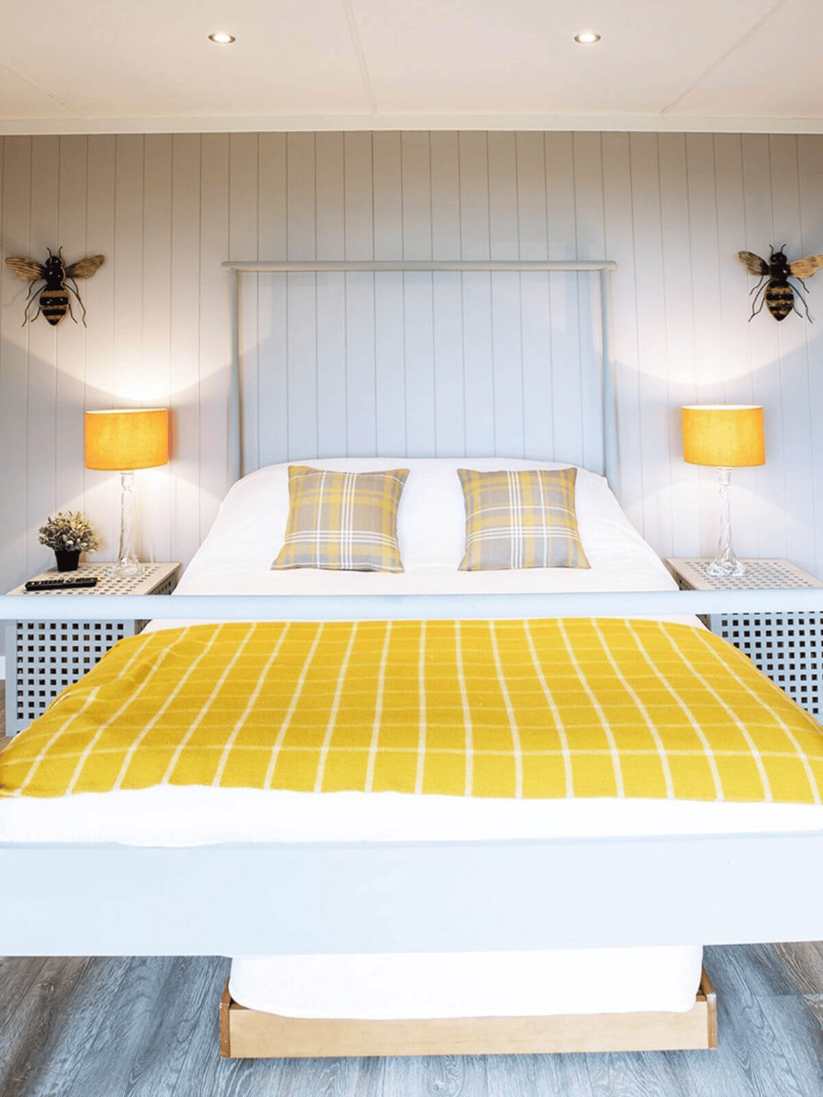 inside view of glamping lodge with double bed and spotlights and yellow checked blanket and checked cushions and bumble bee wall hangings and grey flooring and yellow lamp shades