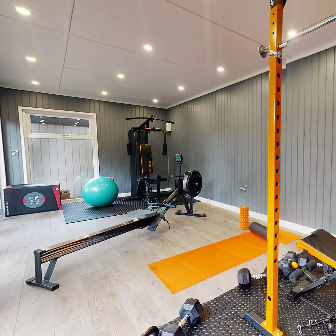 Large garden gym timber building with orange mat, rowing machine, large blue yoga ball and squat rack with dumbbells on floor in front. 