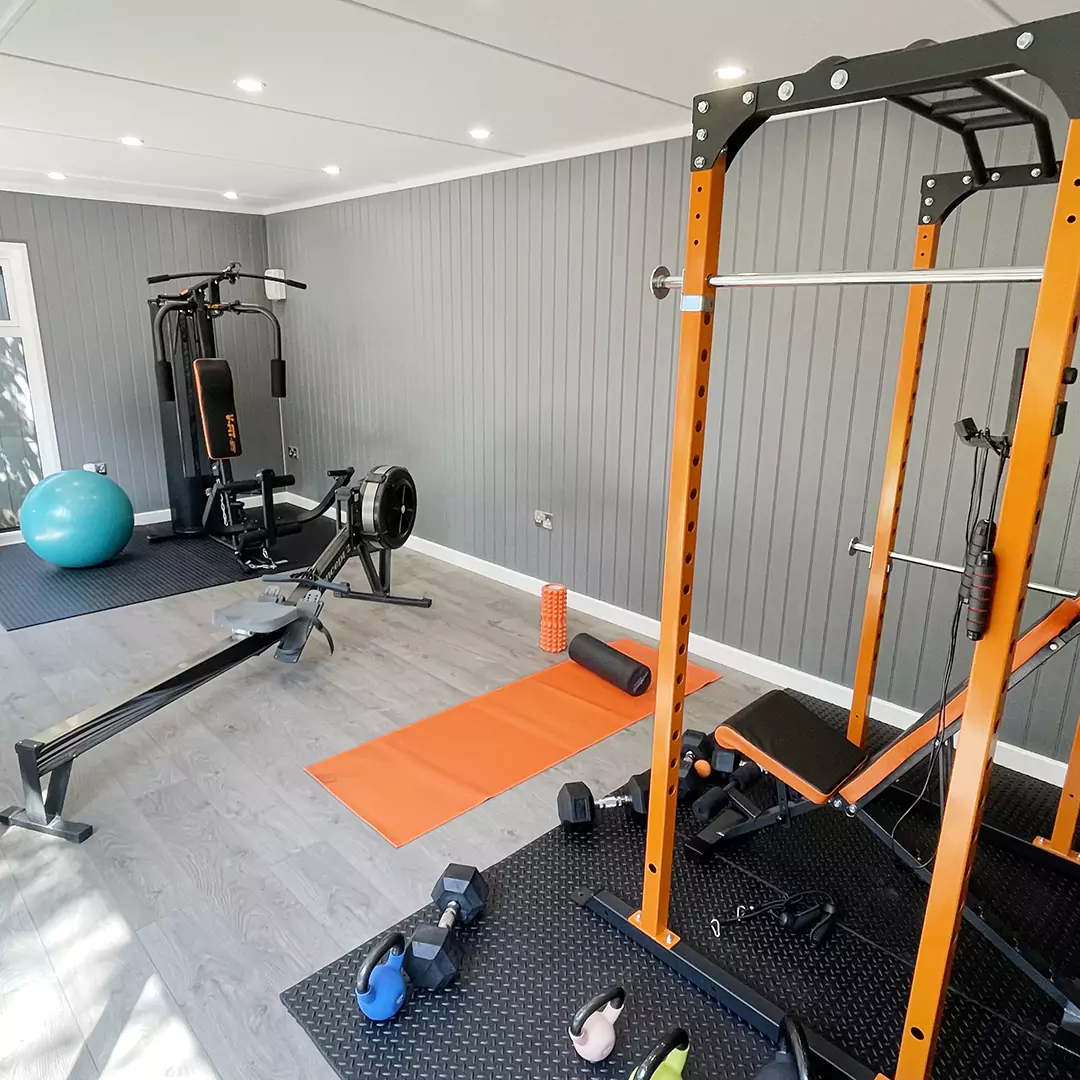 large glass doors inside garden gym building with large orange mat, body roller, squat rack and dumbbells