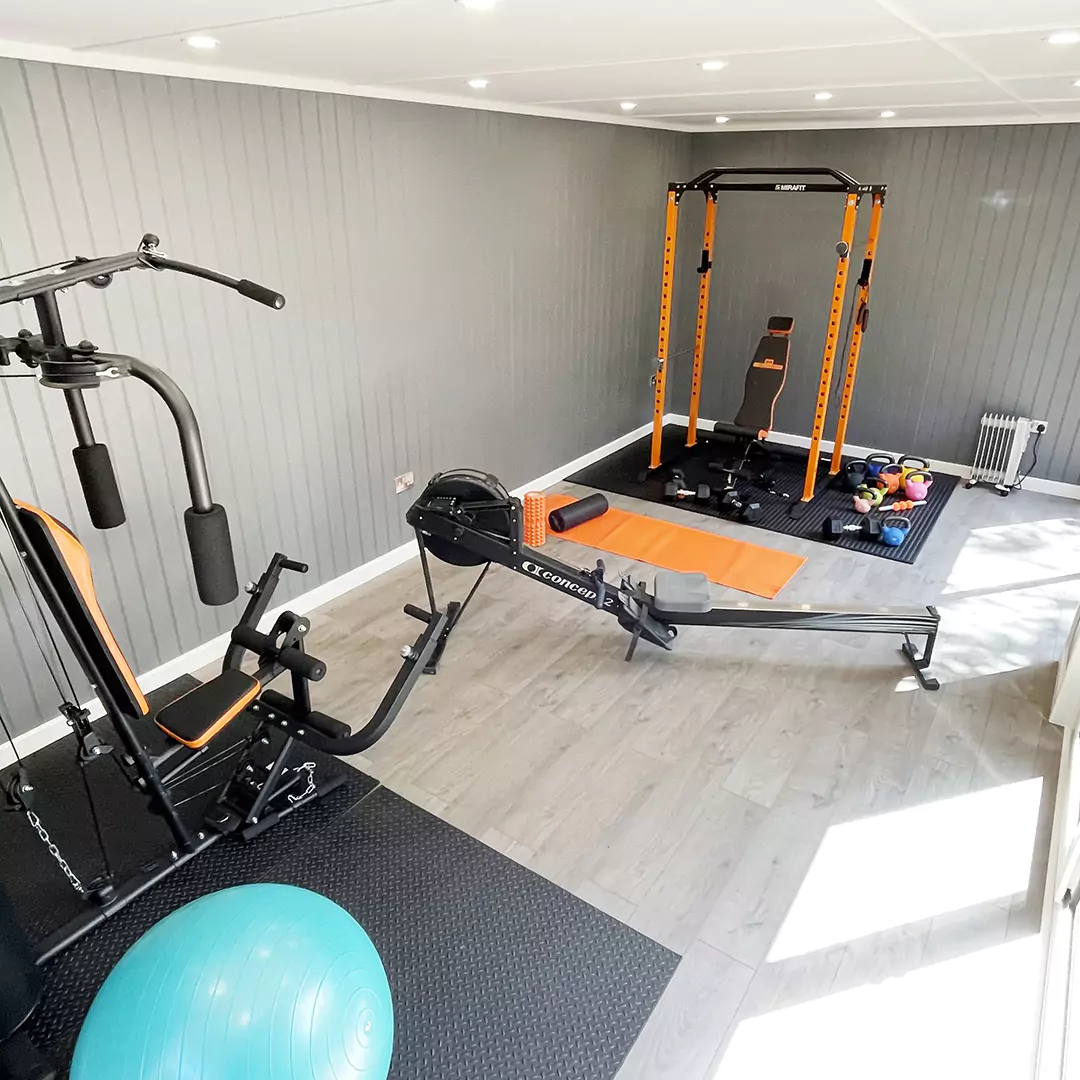 large grey walls inside garden gym building with large orange squat rack, orang mat, rowing machine, yoga ball and upper body machine