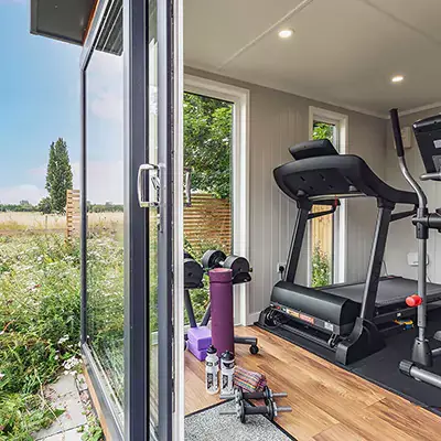 Garden Gym Building with sliding doors and view of running machine, dumbbells, mat and body roller with garden grass and flowers in background