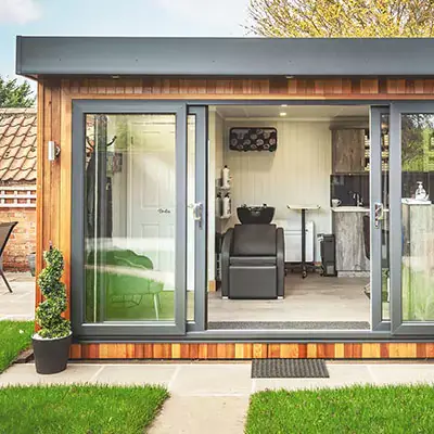 Cabin Master Hairdresser Studio in garden with patio pathway and grassy area and potted plants and sliding doors with internal view of room with black leather chair  