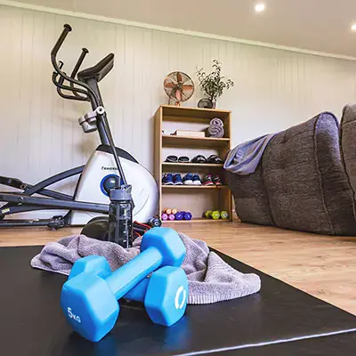 Garden Gym Building Internal shot with dumbbells in foreground and mat and towel and water bottle and dumbbell rack with elliptical machine and grey sofas